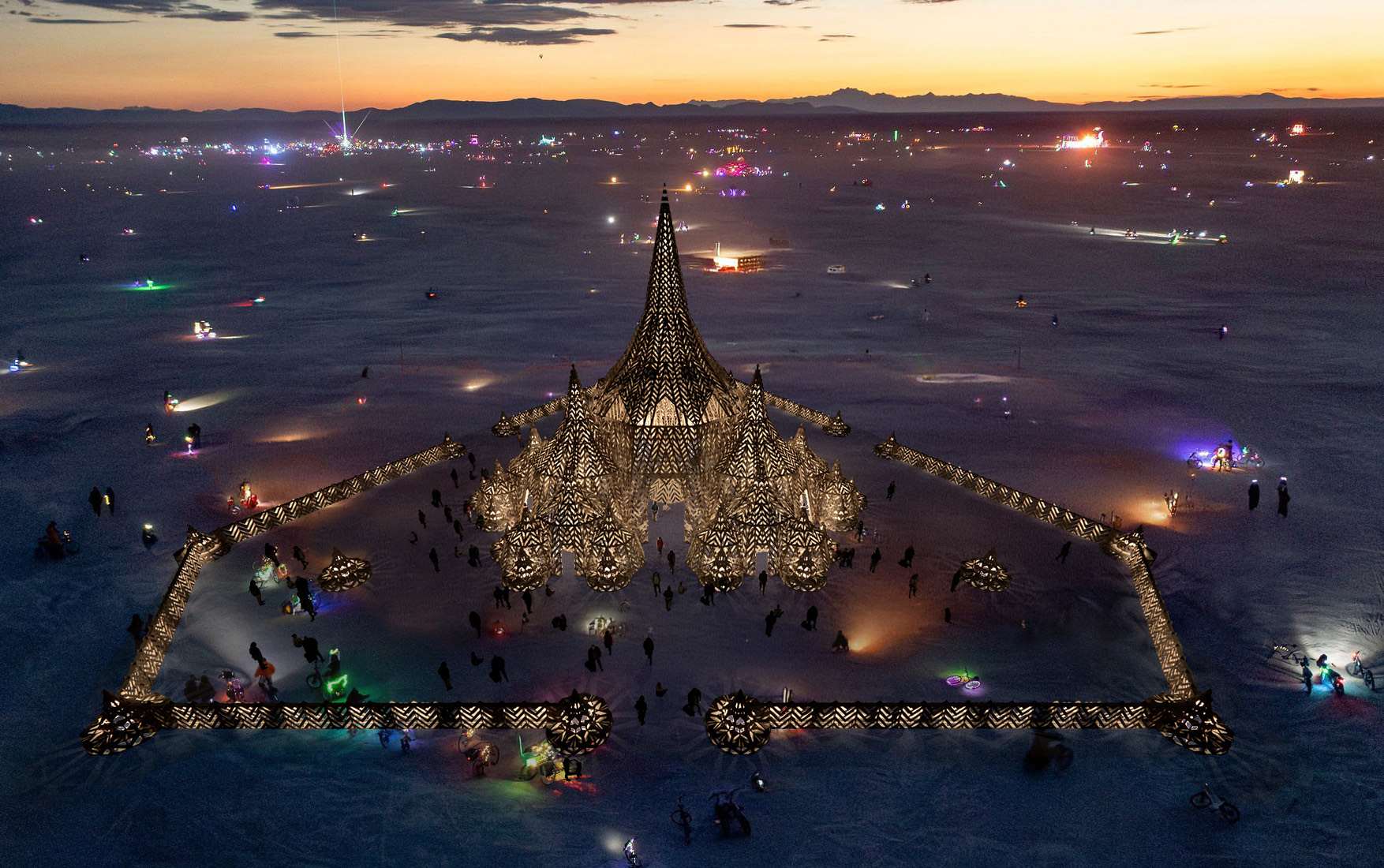 Aerial view of The Temple 2027 at dusk: the lit timber spires and courtyard wall glowing on the playa, with Black Rock City's lights behind